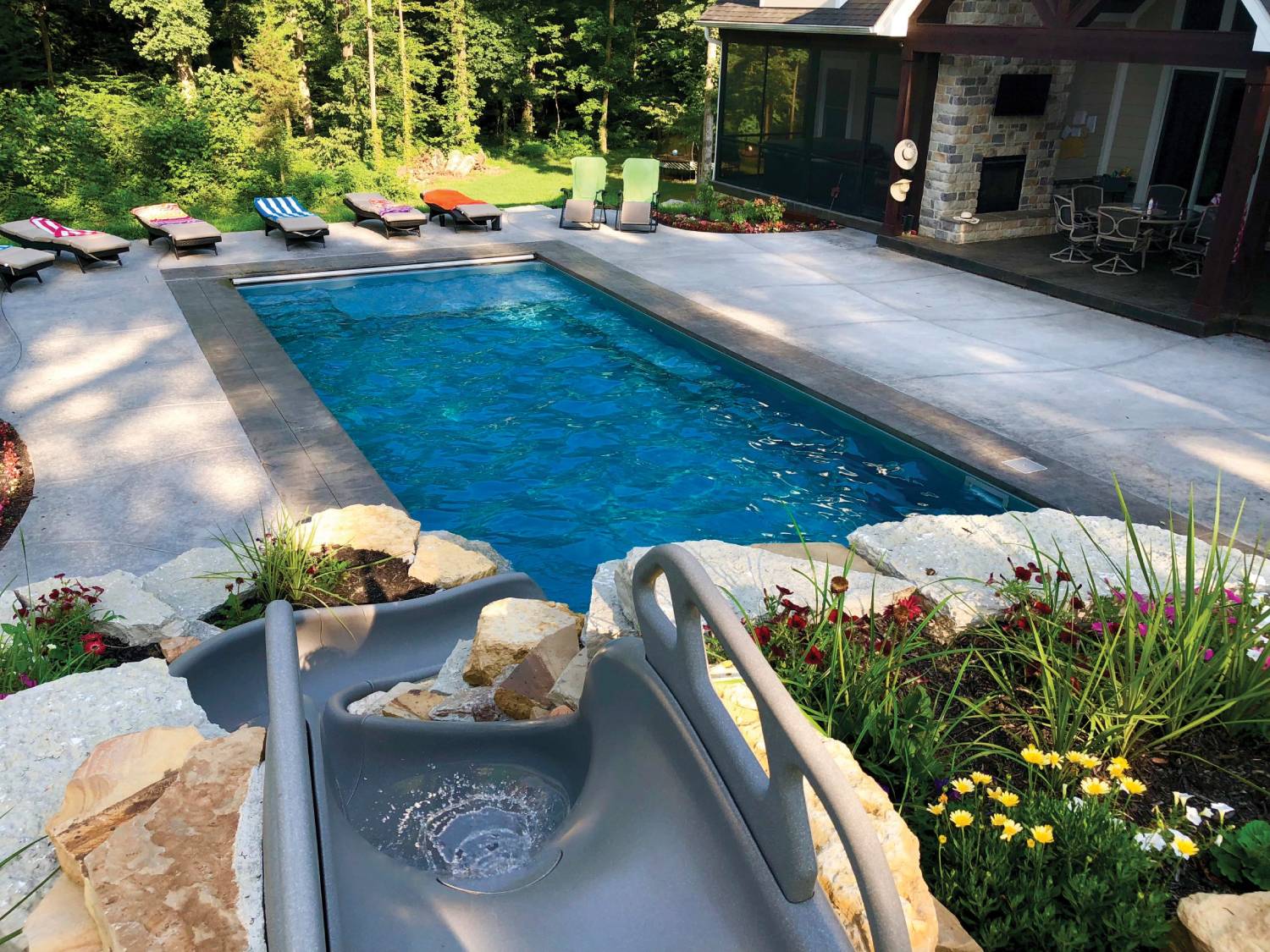 Built-in rectangular pool behind a home, with concrete patio surrounding, and deck chairs along the far side. Pool slide in foreground.